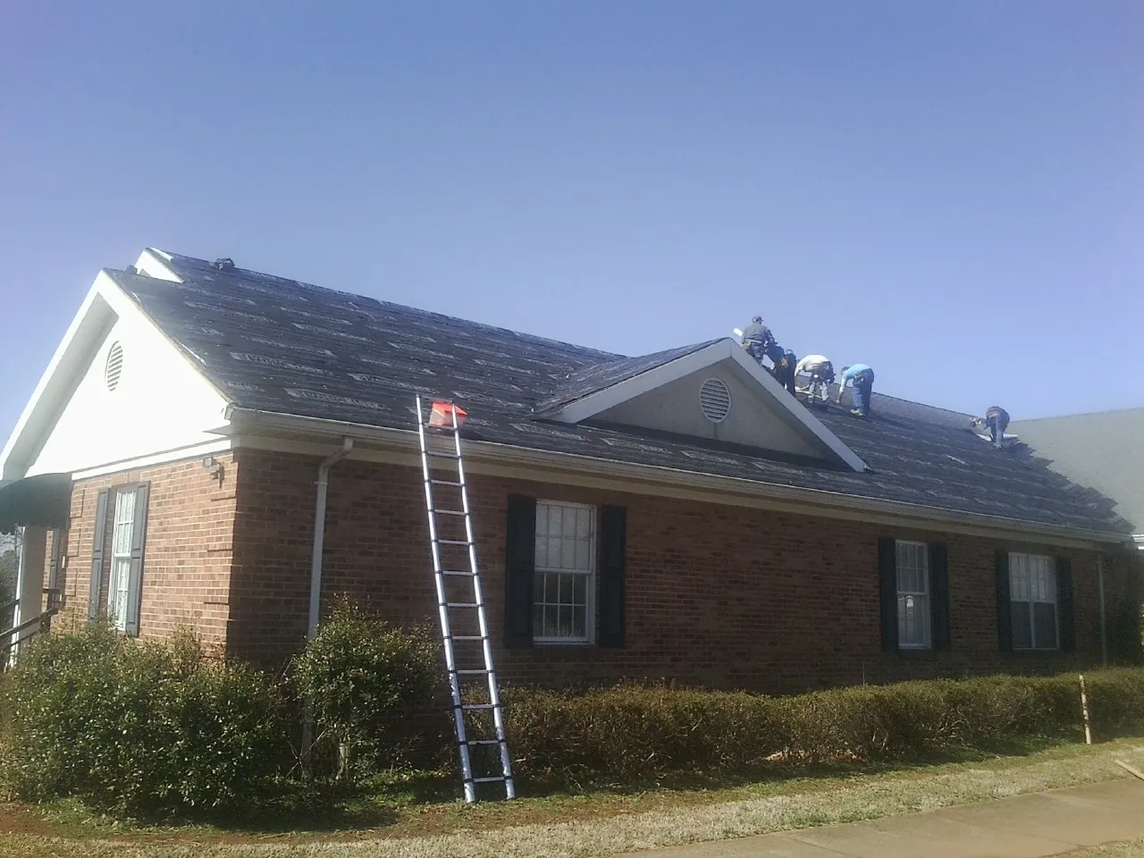 Crew tearing shingles off a brick ranch roof with a ladder set — Emergency Roof Repair work by our crew in Clyde